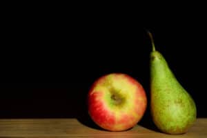 Ein roter und ein grüner Apfel liegen neben einer grünen Birne auf einer Holzoberfläche vor einem schwarzen Hintergrund und werden von der Seite beleuchtet, um ihre Texturen hervorzuheben.