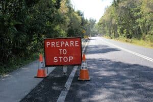 Eine Straße mit einem „Prepare to Stop“-Schild und zwei „Men at Work“-Kegeln am Straßenrand. Die Straße ist von Bäumen und Grünflächen gesäumt, und die Szene lässt darauf schließen, dass eine Baustelle oder Straßenbauarbeiten bevorstehen.