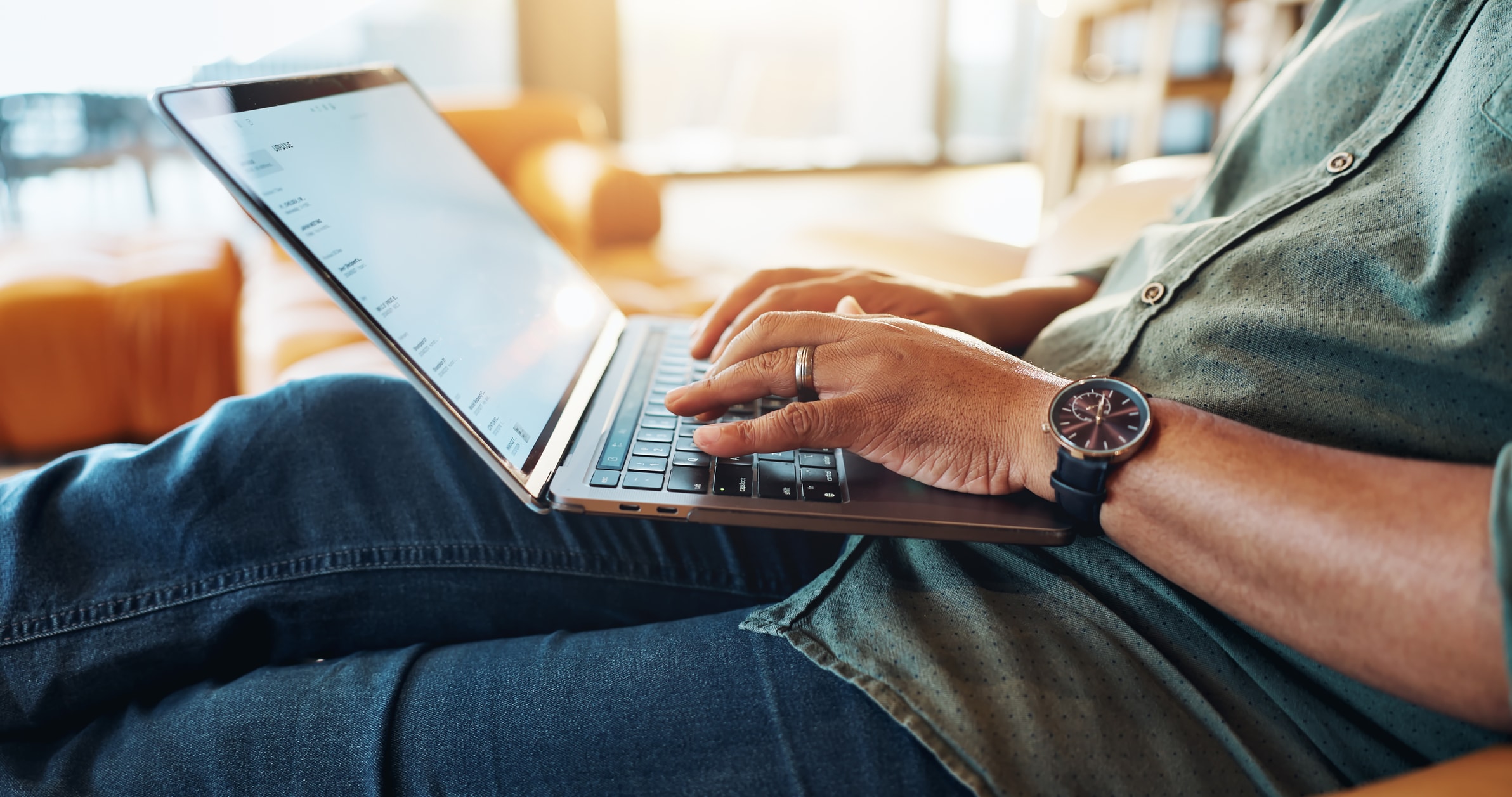 Eine Person sitzt mit einem Laptop auf einem Sofa und tippt mit beiden Händen. Sie trägt eine Armbanduhr und blaue Jeans. Vom Hintergrund fällt Sonnenlicht herein.