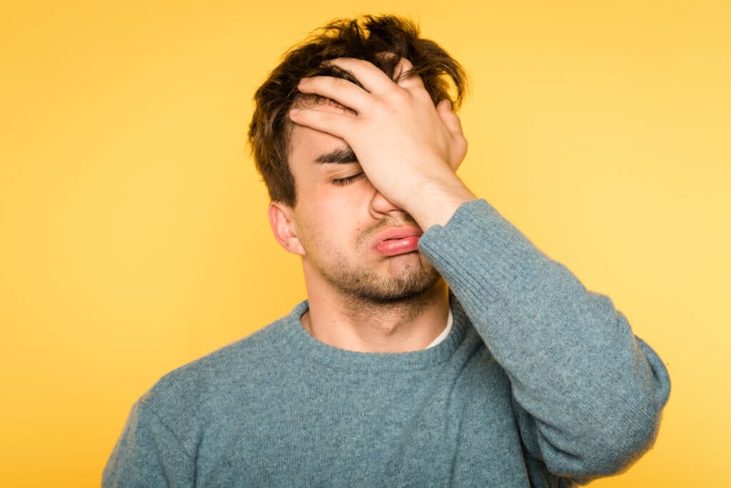 Ein Mann in einem blauen Pullover steht vor einem gelben Hintergrund, bedeckt sein Gesicht mit der Hand, runzelt die Stirn und scheint frustriert oder verärgert zu sein.