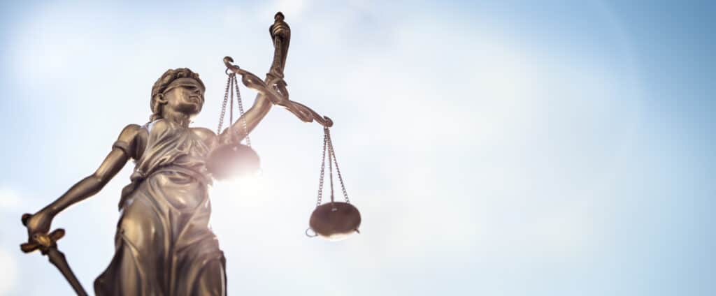 Bronzestatue von Lady Justice, die eine Waage und ein Schwert vor einem hellen Himmelshintergrund hält, als Symbol für Recht und Gerechtigkeit.