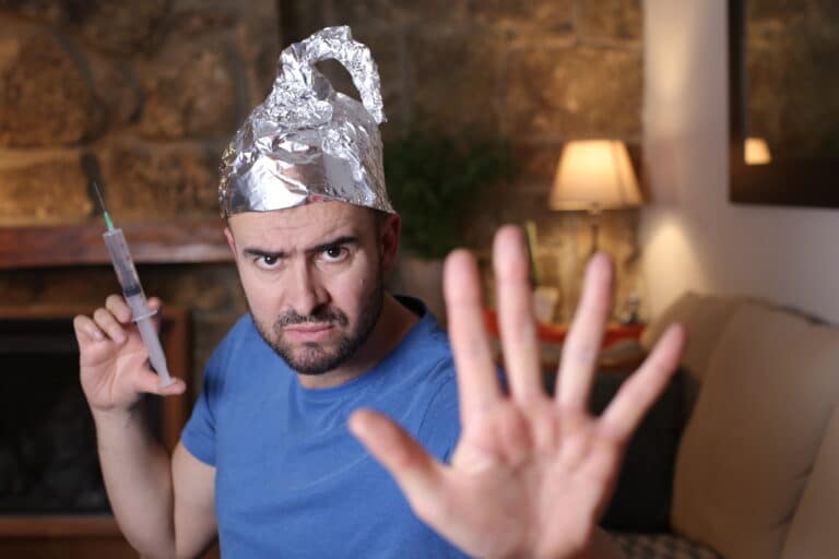 Ein Mann mit einem Hut aus Alufolie hält eine Spritze in einer Hand und streckt die andere Hand nach vorne, als wolle er "Stopp" signalisieren. Er sieht ernst aus und sitzt in einem gemütlichen Wohnzimmer mit einer Lampe und einer Steinmauer im Hintergrund.