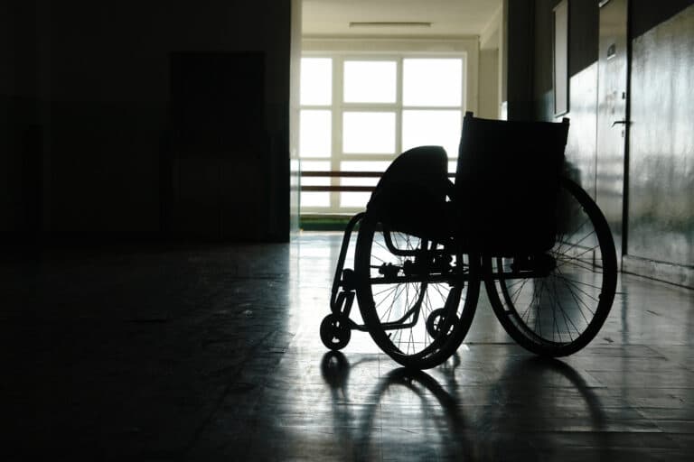 Ein einsamer Rollstuhl steht in einem schwach beleuchteten Flur, durch dessen große Fenster im Hintergrund Sonnenlicht fällt und Schatten auf den Boden wirft.