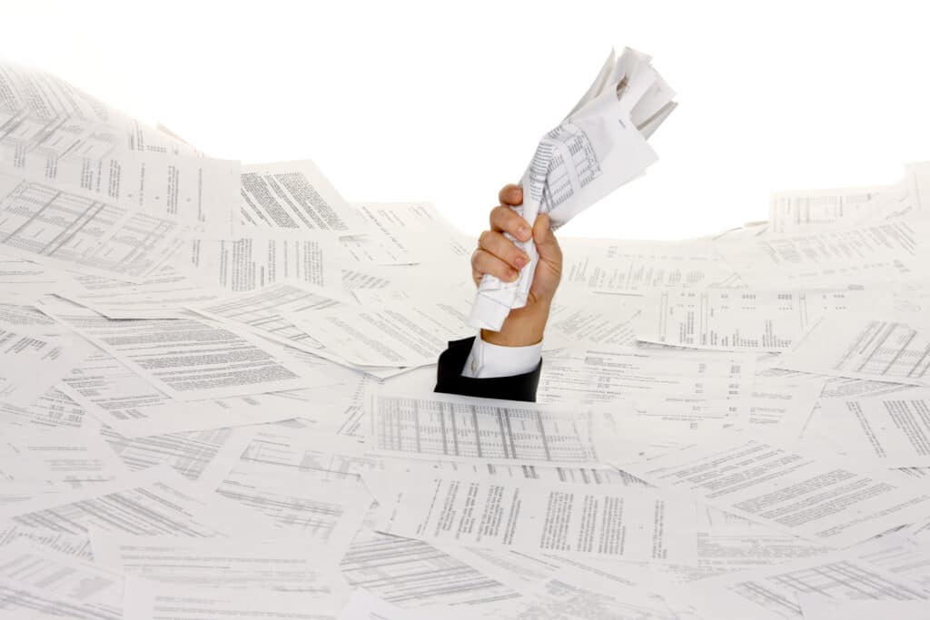 Die Hand einer Person taucht aus einem riesigen Stapel Papierkram auf und greift nach einem Bündel von Dokumenten, was darauf hindeutet, dass sie von Papierkram oder Verwaltungsaufgaben überwältigt ist.