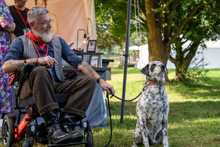 Ein Mann im Rollstuhl, der eine Brille und ein rotes Halstuch trägt, lächelt, während er die Leine eines gefleckten Hundes hält, der neben ihm im Freien in der Nähe eines Zeltes und von Bäumen sitzt.