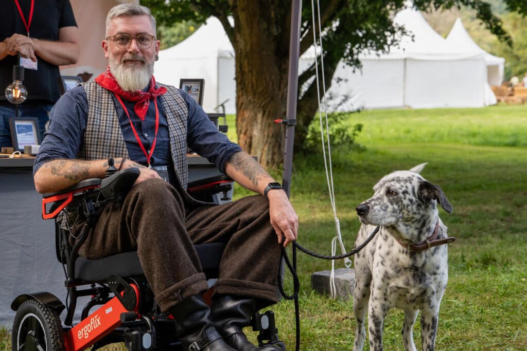 Ein bärtiger Mann im Rollstuhl, der eine Brille, einen roten Schal und eine Tweedweste trägt, sitzt im Freien und hält einen gefleckten Hund an der Leine. Im Hintergrund sind weiße Zelte und grünes Gras zu sehen.