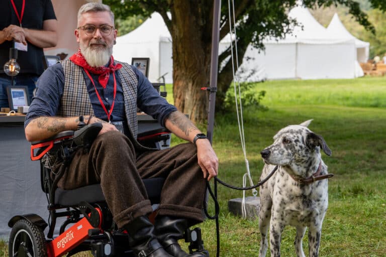 Ein bärtiger Mann im Rollstuhl, der eine Brille, einen roten Schal und eine Tweedweste trägt, sitzt im Freien und hält einen gefleckten Hund an der Leine. Im Hintergrund sind weiße Zelte und grünes Gras zu sehen.