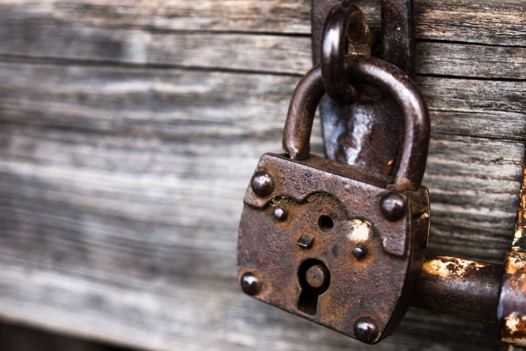 Eine Nahaufnahme eines rostigen Vorhängeschlosses, das an einer verwitterten Holztür hängt und Anzeichen von Alter und Abnutzung aufweist. Die dunkle Metalloberfläche des Schlosses ist verfärbt und von Rost zerfressen.