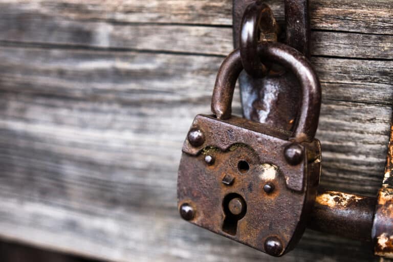 Eine Nahaufnahme eines rostigen Vorhängeschlosses, das an einer verwitterten Holztür hängt und Anzeichen von Alter und Abnutzung aufweist. Die dunkle Metalloberfläche des Schlosses ist verfärbt und von Rost zerfressen.