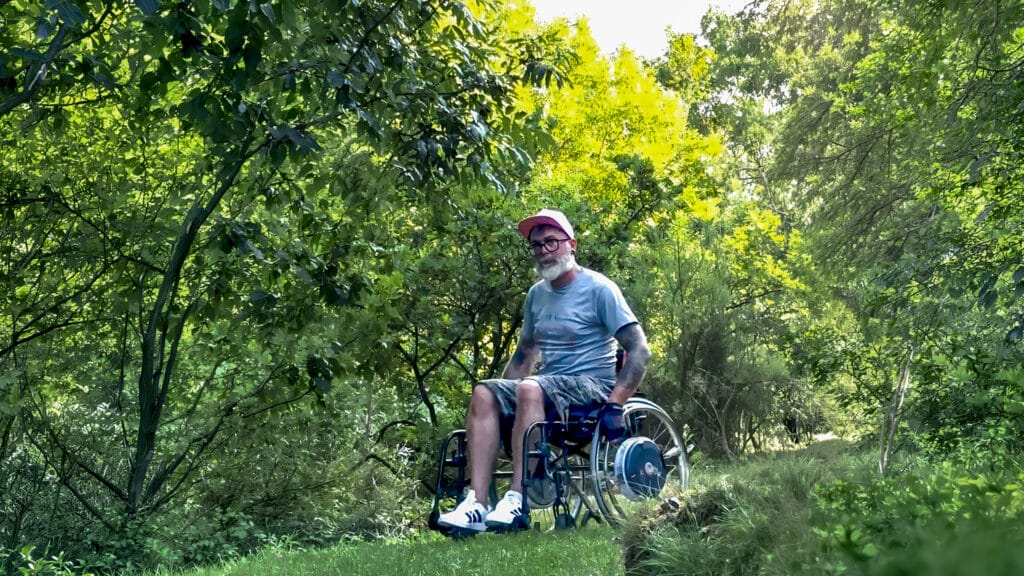 Ein Mann im Rollstuhl, der eine rosa Mütze, ein graues T-Shirt und Shorts trägt, befindet sich draußen auf einem grasbewachsenen Weg, umgeben von üppig grünen Bäumen und Laub, und genießt einen sonnigen Tag.