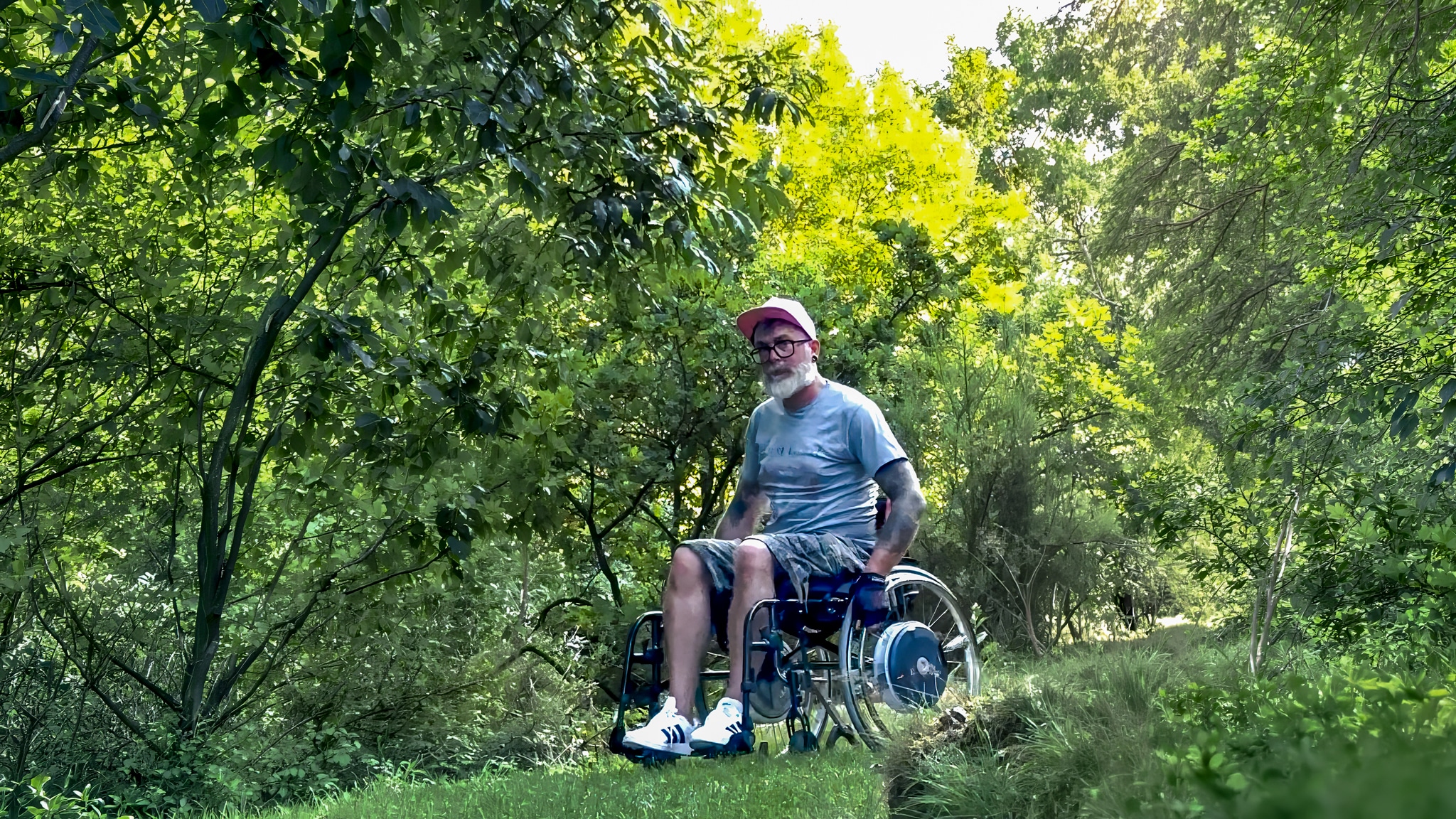 Ein Mann im Rollstuhl, der eine rosa Mütze, ein graues T-Shirt und Shorts trägt, befindet sich draußen auf einem grasbewachsenen Weg, umgeben von üppig grünen Bäumen und Laub, und genießt einen sonnigen Tag.