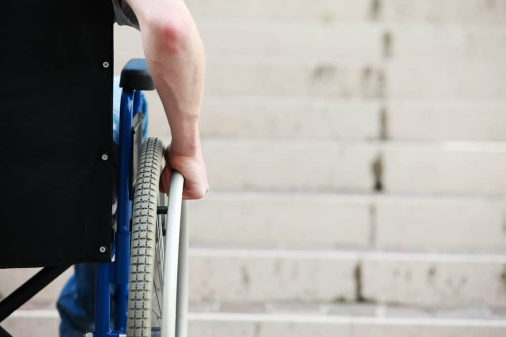 Eine Person im Rollstuhl steht vor einer Treppe aus Beton, hält sich am Rad fest und blickt zu dem Hindernis hinauf, wodurch die Herausforderungen im Bereich der Barrierefreiheit deutlich werden.